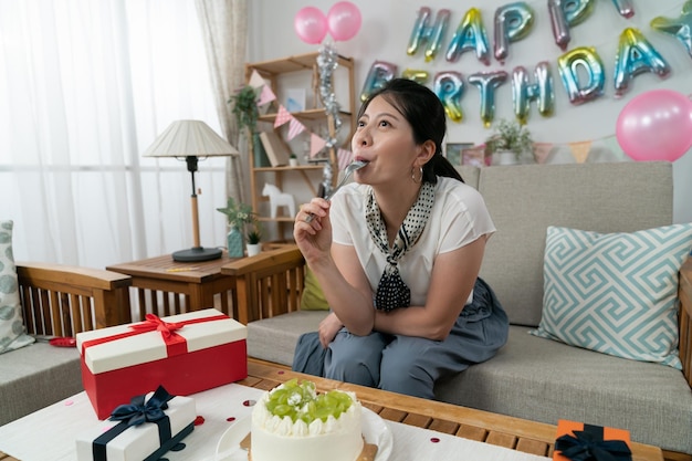 gelukkige aziatische vrouw die room likt en lepel in de mond houdt terwijl ze alleen geniet van smakelijke cake op haar verjaardagsfeestje in het appartement met feestdecors.