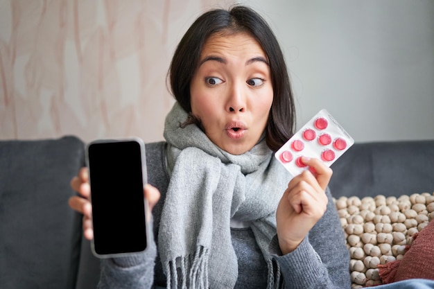 Gelukkige aziatische vrouw die verkouden wordt met smartphonescherm en pillen die online dokter app g aanbevelen