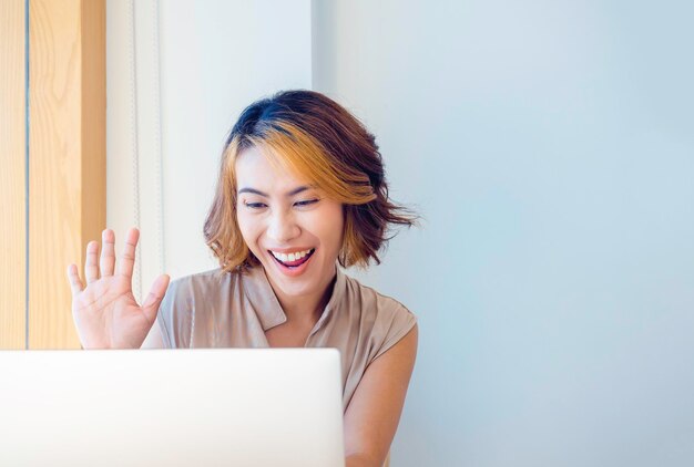 Gelukkige Aziatische vrouw met kort haar zegt hallo met haar hand omhoog tijdens het gebruik van een laptopcomputer, videogesprek op een witte muurachtergrond met kopieerruimte. Aantrekkelijke volwassen vrouwelijke facetime, videoconferentie thuis.