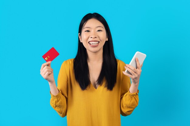 Gelukkige aziatische vrouw met smartphone en bankkaart op blauw