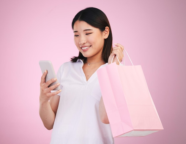 Gelukkige Aziatische vrouw telefoon en boodschappentas in betalingsaankoop of kopen tegen een roze studio achtergrond Vrouwelijke persoon of shopper op mobiele smartphone bankieren app in e-commerce verkoop of korting