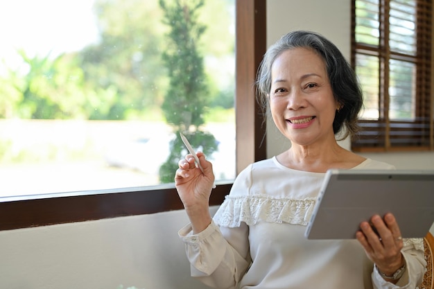 Gelukkige Aziatische vrouw uit de jaren 60 die bij het raam in haar woonkamer zit en een tablet gebruikt