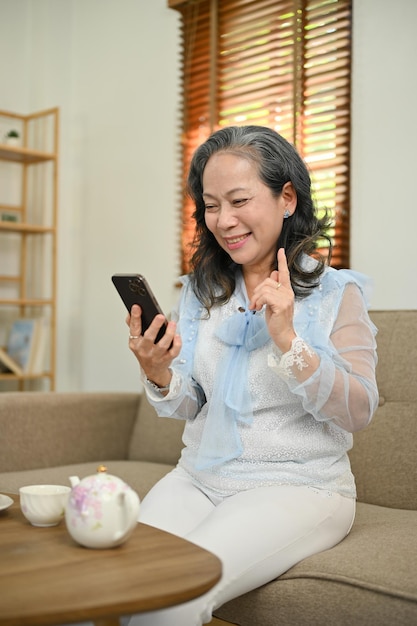 Gelukkige Aziatische vrouw uit de jaren 60 die haar smartphone gebruikt om een videogesprek te voeren met haar kleinkind