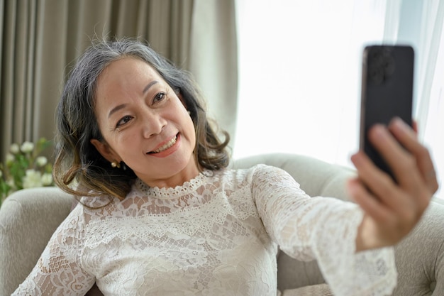 Gelukkige Aziatische vrouw van middelbare leeftijd gebruikt haar smartphone om een selfie te maken in haar woonkamer