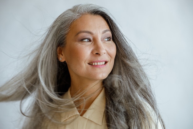 Foto gelukkige aziatische vrouw van middelbare leeftijd met vliegende grijze sloten staat op een lichte achtergrond