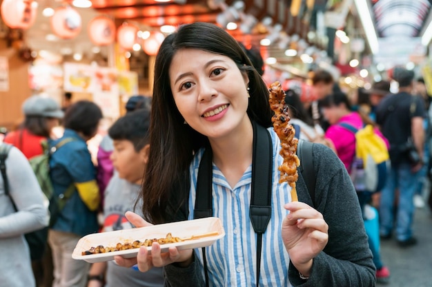 gelukkige Aziatische vrouwelijke reiziger glimlachend in de camera terwijl u geniet van heerlijk gegrild varkensvlees op stok in de drukke kuromon ichiba-markt in Osaka, japan