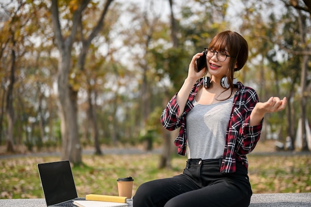 Gelukkige Aziatische vrouwelijke student die aan de telefoon praat terwijl ze ontspant in het campuspark