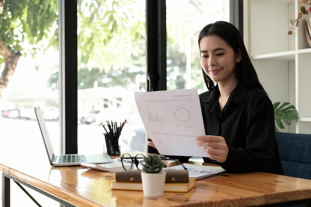 Gelukkige Aziatische zakenvrouw die werkt met papierwerkgegevens, grafiekdocumenten en laptopcomputerzaken