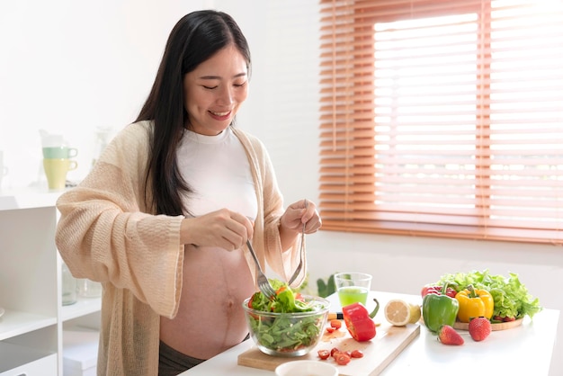 Gelukkige aziatische zwangere vrouw die salade thuis kookt die verse groene salade doet