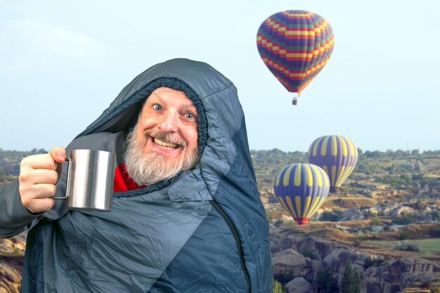 Gelukkige baarde man in een slaapzak met een kop koffie tegen de achtergrond van de natuur