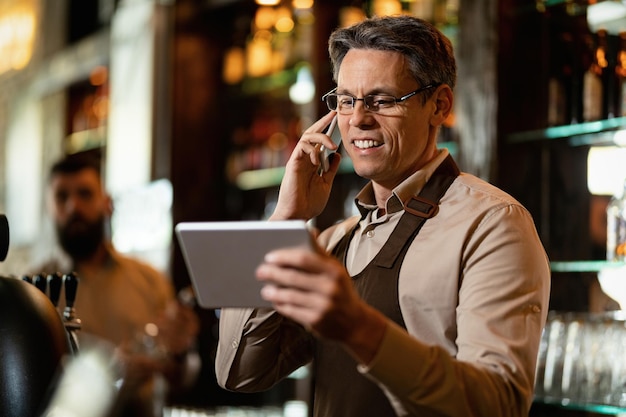 Gelukkige barman die touchpad gebruikt tijdens het communiceren op mobiele telefoon in een bar.