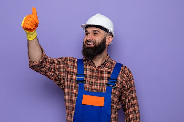 Gelukkige, bebaarde bouwman in bouwuniform en veiligheidshelm met rubberen handschoenen die omhoog lacht en vrolijk duimen laat zien
