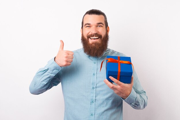 Gelukkige bebaarde man toont duim terwijl hij een geschenkdoos vasthoudt.