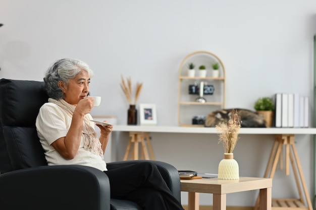 Gelukkige bejaarde vrouw ontspant zich op een fauteuil en drinkt thee geniet van stressvrije vreedzame stemming