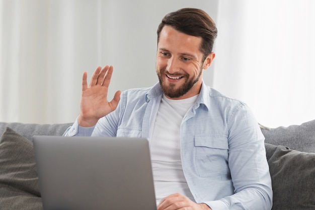 Gelukkige blanke man gebruikt laptopcomputer voor videogesprekken terwijl hij thuis op de bank zit, glimlachende blanke man chat met vrienden of familie via videoconferentie op sociale netwerken, begroetingsgebaar