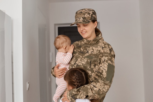 Gelukkige blije militaire moeder die camouflage-uniform en hoed draagt, haar kinderen knuffelt en naar kinderen kijkt met een aangename glimlach, liefde uitdrukkend, geserveerd in het leger.