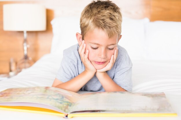 Gelukkige blonde jongen die op bed ligt dat een verhalenboek leest