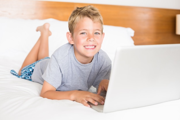 Gelukkige blonde jongen die op bed ligt dat laptop met behulp van