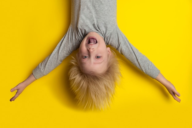 Gelukkige blonde jongen opknoping ondersteboven met uitgestrekte armen. Kind op heldere gele achtergrond.