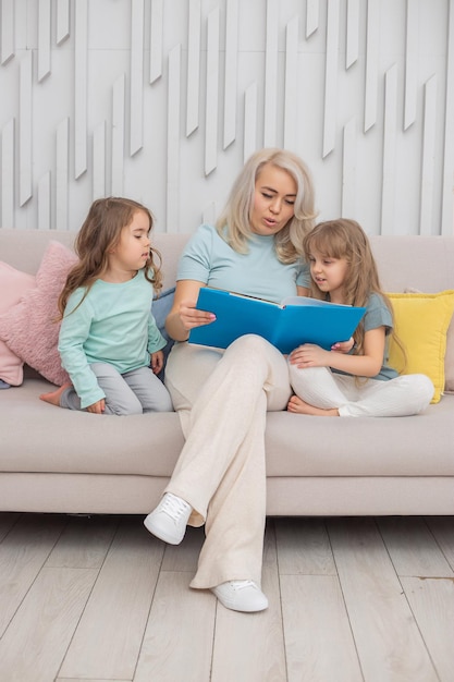 Gelukkige blonde moeder leest een boek voor aan haar schattige dochters die op de bank in de woonkamer zitten. gelukkig familieconcept.