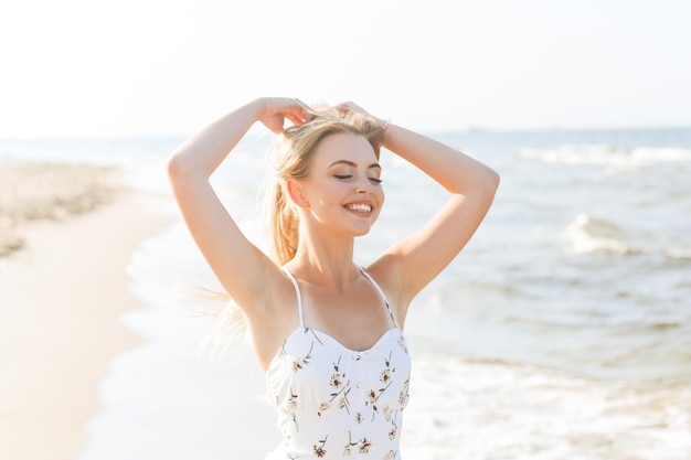 Gelukkige blonde mooie vrouw op het oceaanstrand die in een witte zomerjurk staat en handen opheft.