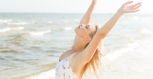 Gelukkige blonde mooie vrouw op het oceaanstrand in een witte zomerjurk, handen omhoog.