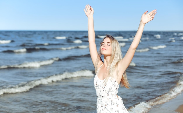Gelukkige blonde mooie vrouw op het strand van de oceaan, staande in een witte zomerjurk, handen opstekend.