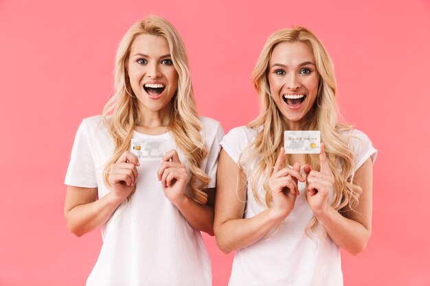 Foto gelukkige blonde tweeling die in t-shirts draagt die creditcards houdt en de voorzijde over roze muur bekijkt