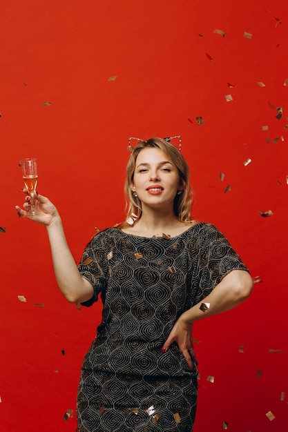 Gelukkige blonde vrouw in glanzende mode kleding is blij met glas champagne en poseren met confetti op rode achtergrond