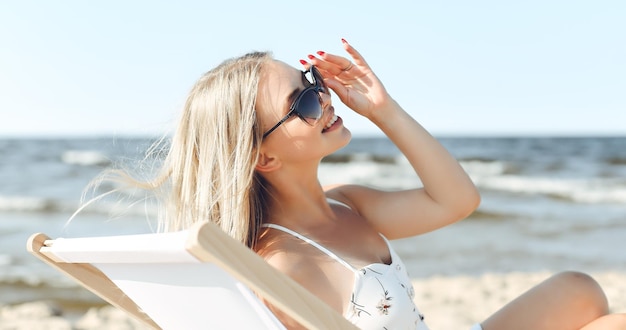 Gelukkige blonde vrouw met een zonnebril en ontspannen op een houten leunstoel op het strand