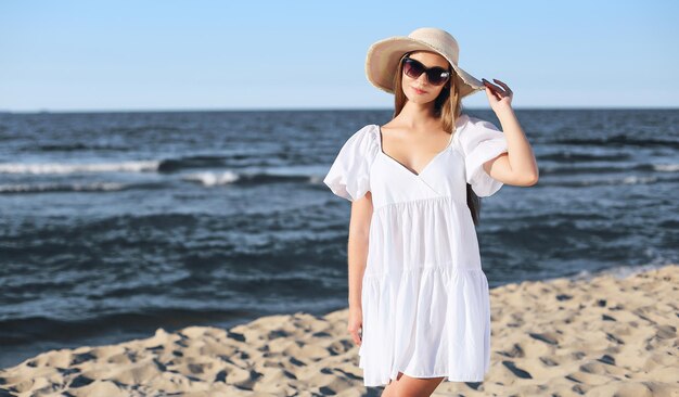 Gelukkige blonde vrouw poseert op het oceaanstrand met zonnebril en een hoed Avondzon