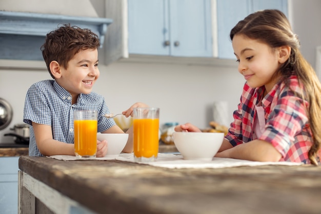 Gelukkige broers en zussen. Knappe vrolijke donkerharige jongetje kijken naar zijn zus en glimlachen en ze hebben gezond ontbijt