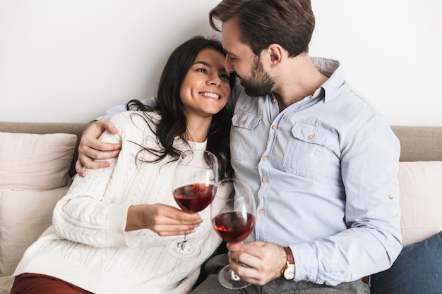 gelukkige brunette paar man en vrouw die samen wijn uit een bril drinken terwijl ze thuis op de bank in een lichte kamer zitten