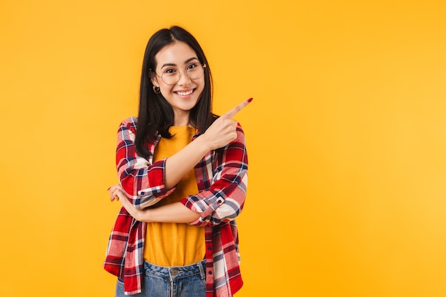 Gelukkige brunette vrouw in bril glimlachend terwijl ze met de vinger opzij wijst geïsoleerd over gele muur