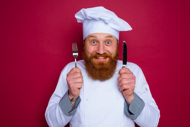 Gelukkige chef-kok met baard en rode schort houdt bestek in de hand