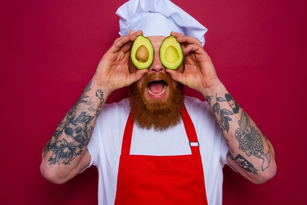 Gelukkige chef-kok met baard en rood schort houdt een avocado vast