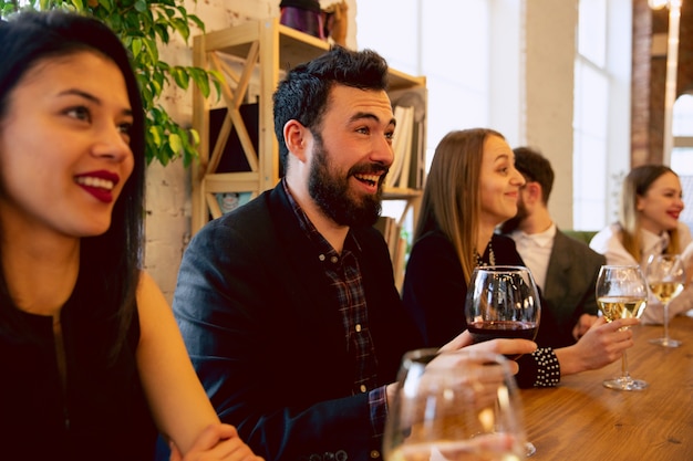 Gelukkige collega's vieren tijdens bedrijfsfeest, bedrijfsevenement. Jonge blanke mensen in zakelijke kleding praten, wijn drinken. Concept van kantoorcultuur, teamwork, vriendschap, vakantie, weekend.