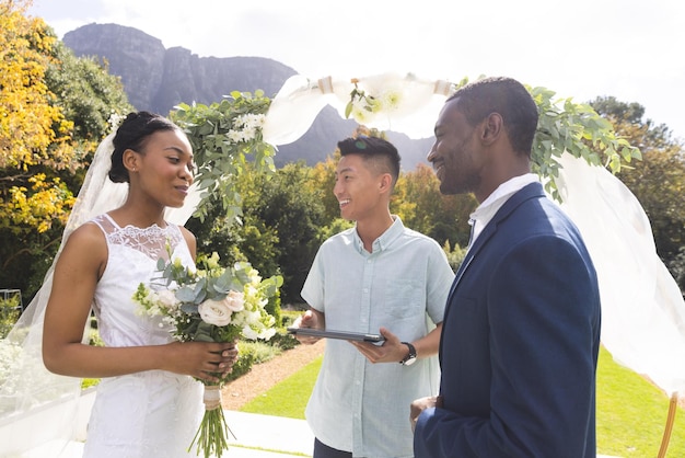 Gelukkige diverse mannelijke officiant met bruid en bruidegom bij een huwelijksceremonie in de buitenlucht in een zonnige tuin