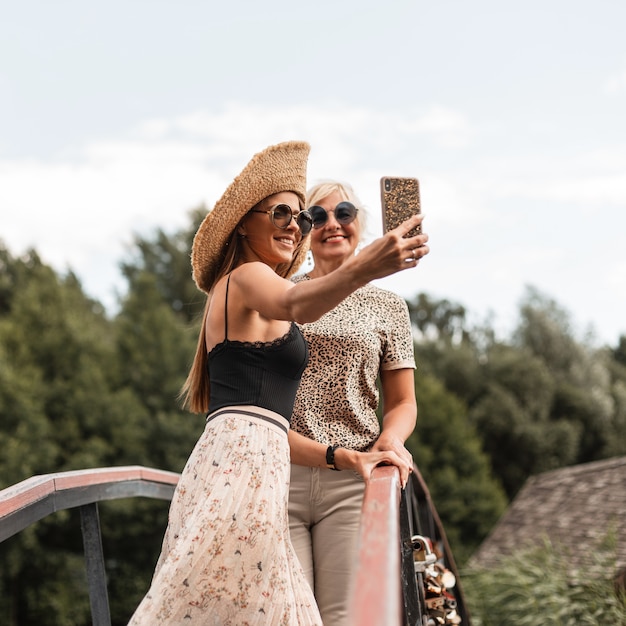 Gelukkige dochter met lachende moeder in modieuze zomeroutfit met strohoed en zonnebril maken een gezamenlijke foto op de smartphone en rust in de natuur van het platteland