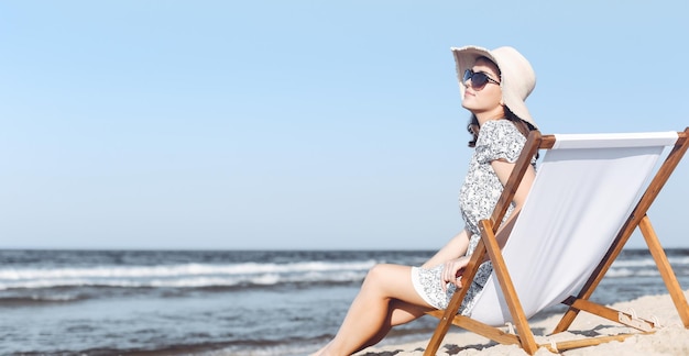 Gelukkige donkerbruine vrouw die zonnebril en hoed draagt die op een houten ligstoel ontspannen bij het oceaanstrand.