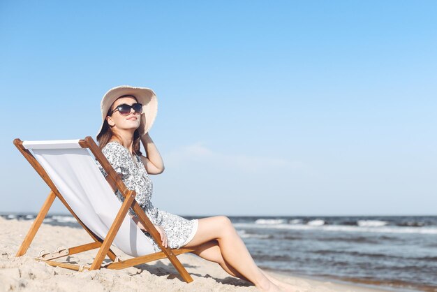 Gelukkige donkerbruine vrouw die zonnebril en hoed draagt die op een houten ligstoel ontspannen bij het oceaanstrand.