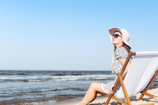 Gelukkige donkerbruine vrouw die zonnebril en hoed draagt die op een houten ligstoel ontspannen bij het oceaanstrand.