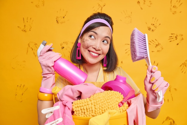 Gelukkige dromerige Aziatische huisvrouw met slordig gezicht glimlacht plasantly houdt spuitwasmiddel en borstel bezig met het schoonmaken van de badkamer en het doen van was poses tegen de gele muur