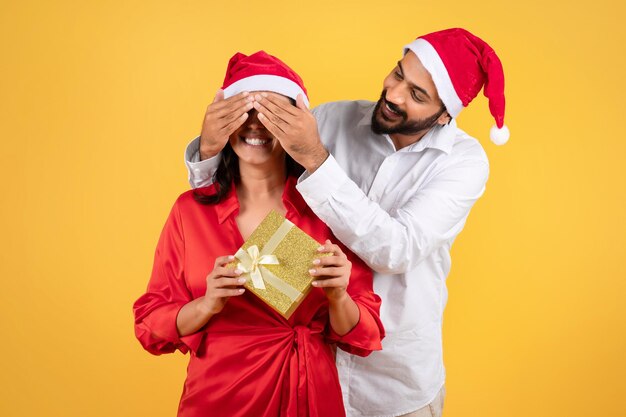 Foto gelukkige duizendjarige arabische man in kerstmuts die de ogen sluit van een vrouw met cadeau geïsoleerd op gele studio