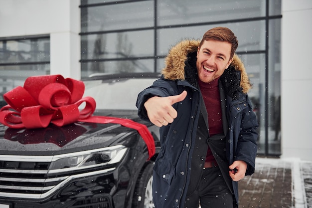 Gelukkige eigenaar van een nieuwe moderne auto die buiten staat en glimlacht. Rode geschenklengte op de auto.