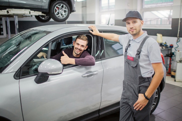 Gelukkige eigenaar zit in de auto en glimlacht. Hij houdt zijn grote duim omhoog. Werknemer staan op auto en houdt zijn hand op het dak van de auto. Ze poseren op camera.