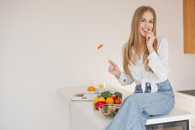 Gelukkige en glimlachende blonde vrouw die bovenop de tafel in de keuken zit en geniet van zelfgemaakte salade tussen kookingrediënten