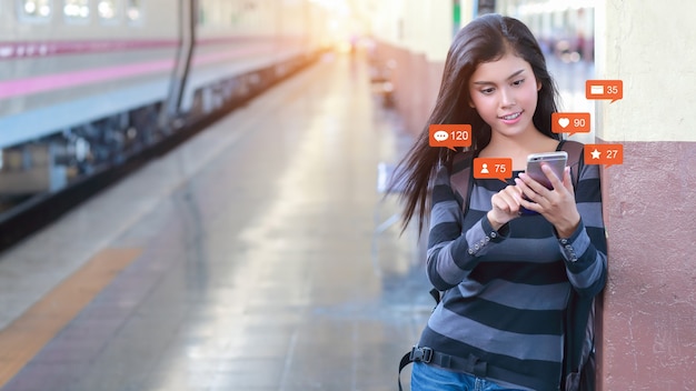 Gelukkige en mooie aziatische vrouw die in het treinstation staat, gebruikt mobiele telefoon met sociale media-interactie, meldingspictogrammen, e-mail, bericht, zoals, liefde, commentaar op het apparaat. Lifestyle met technologie