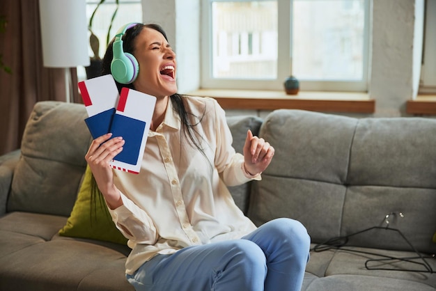 Gelukkige en opgewonden jonge vrouw luistert naar muziek in koptelefoon zit thuis op de bank en toont paspoort met kaartjes