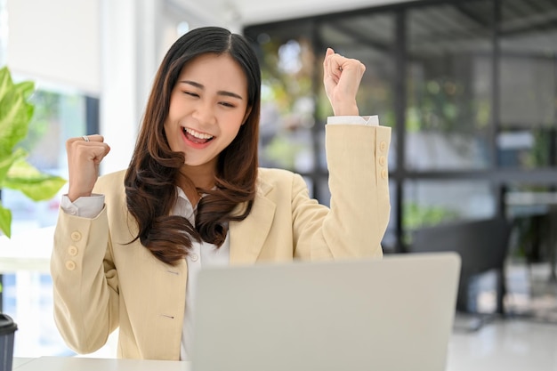 Gelukkige en vrolijke Aziatische zakenvrouw die naar het laptopscherm kijkt en haar succes viert